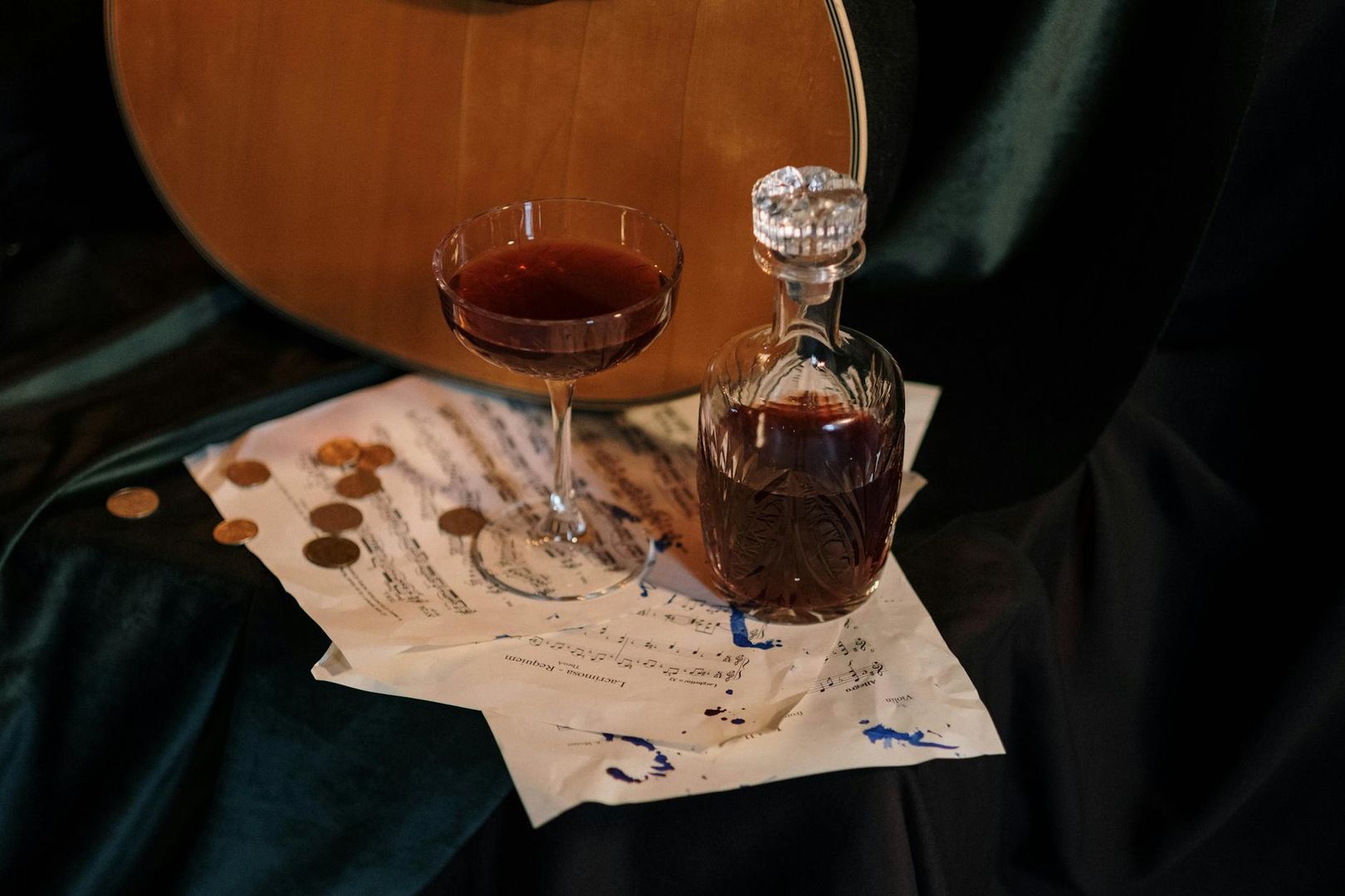Gitarr, notblad, mynt, dryckesglas och karaff på mörkt tyg.
